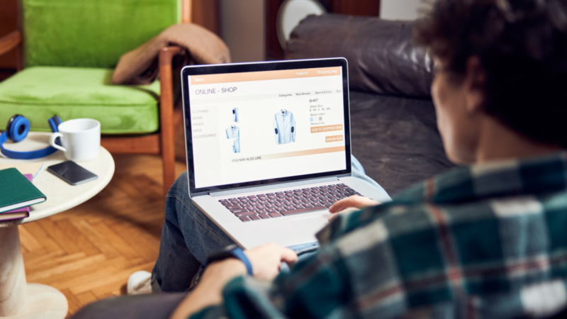 A backshot of a man sitting in a chair with a laptop and shopping online for shirts.