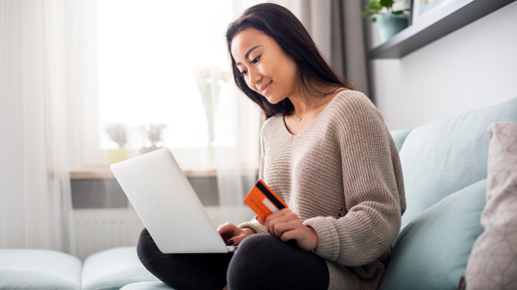 A woman seated on a couch, using a laptop while holding a credit card in her right hand.