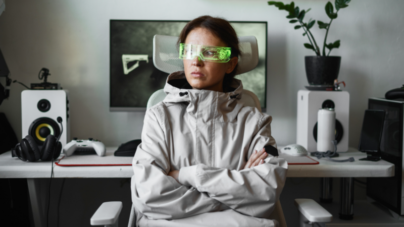 Woman in futuristic green visor, seated with arms crossed. Surrounded by tech, speakers, and a plant in a modern workspace.