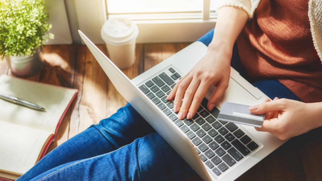 Woman sitting on a wooden floor, using a laptop while holding a credit card in her hand.