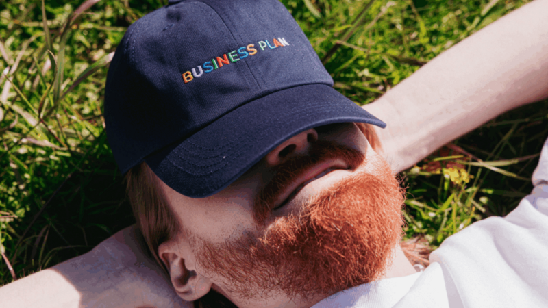 A bearded man lying in the grass, wearing a hat, enjoying a sunny day outdoors.