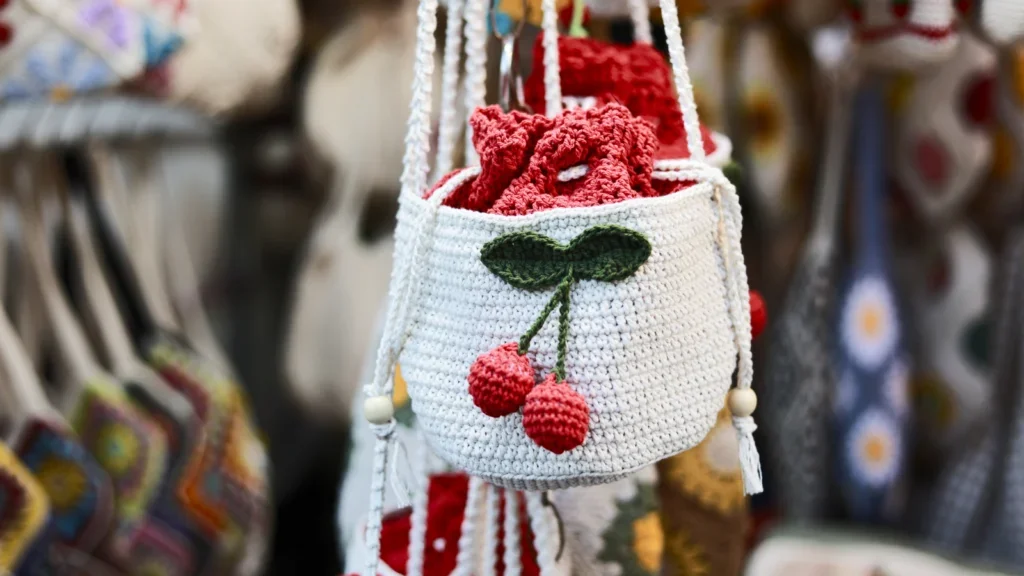 A white crocheted bag with a red cherry design hangs among colorful fabric items.
