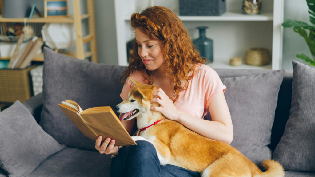 A woman with red hair petting a dog and reading a book.