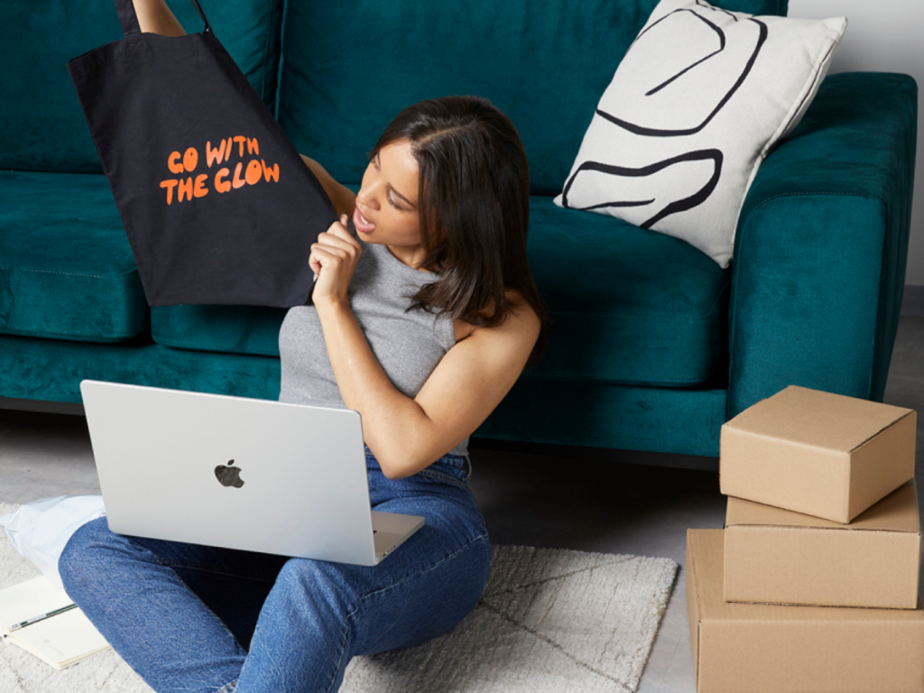 A woman sitting on the floor with a laptop on her lap, holding and looking at a custom tote bag in her hands.