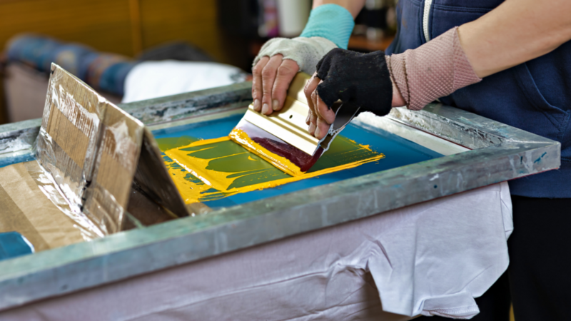 A person is screen printing a white t-shirt using a squeegee with yellow and red ink on a blue screen.