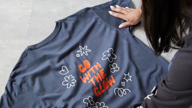 A person is laying down a navy blue shirt with the phrase "GO WITH THE GLOW" and floral designs on it.
