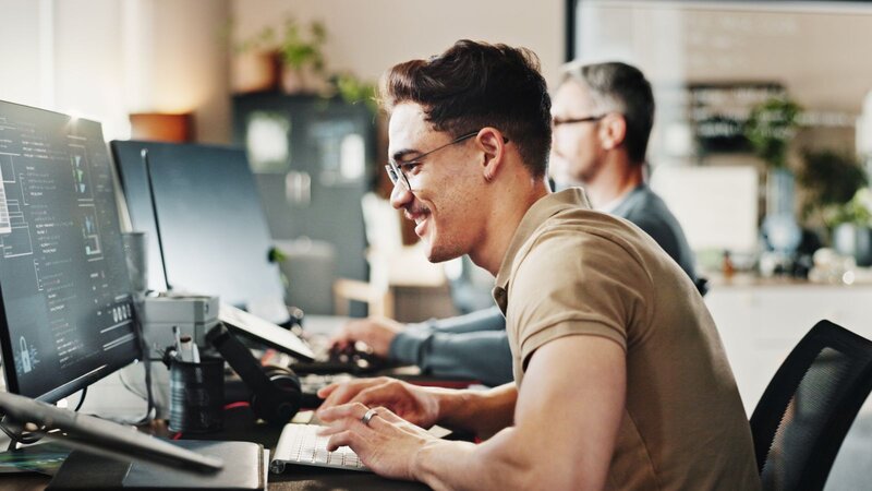 A young man with glasses, sitting at a desk, smiling while working on a computer. In the background, another person is also working at a desk.