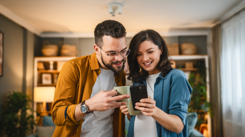 A smiling couple in a cozy living room looks at a smartphone together. The man holds a mug. Shelves in the background.