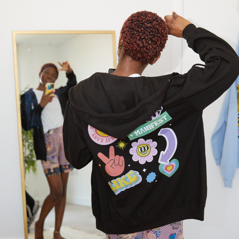 A woman wearing a black hoodie adorned with various colorful prints, standing confidently against a neutral background.