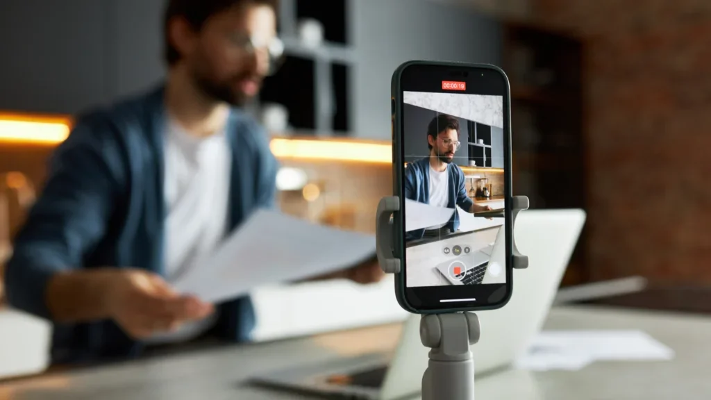 A smartphone mounted on a tripod records a man in a modern kitchen holding papers with a laptop in front of him.