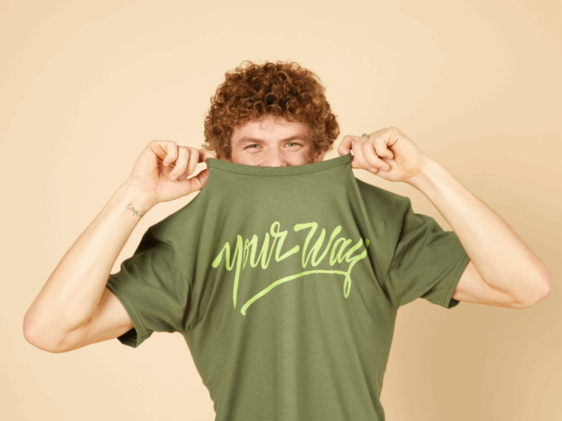 A smiling man playfully lifts their green t-shirt to expose their eyes. The shirt reads "your way" in light green script.