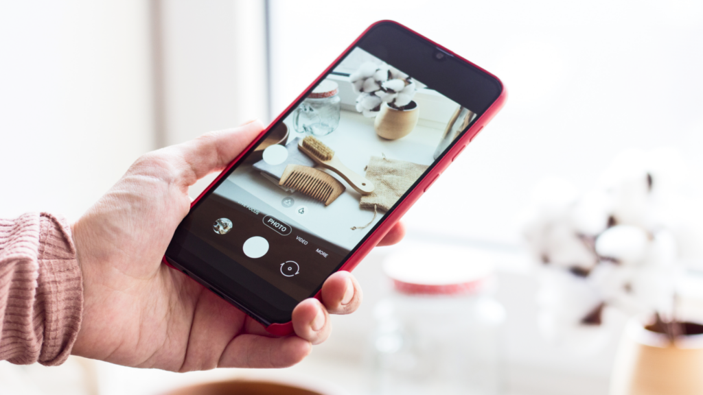 A hand holds a smartphone with a red case, capturing a photo of a wooden comb, jar, and cotton balls on a windowsill.