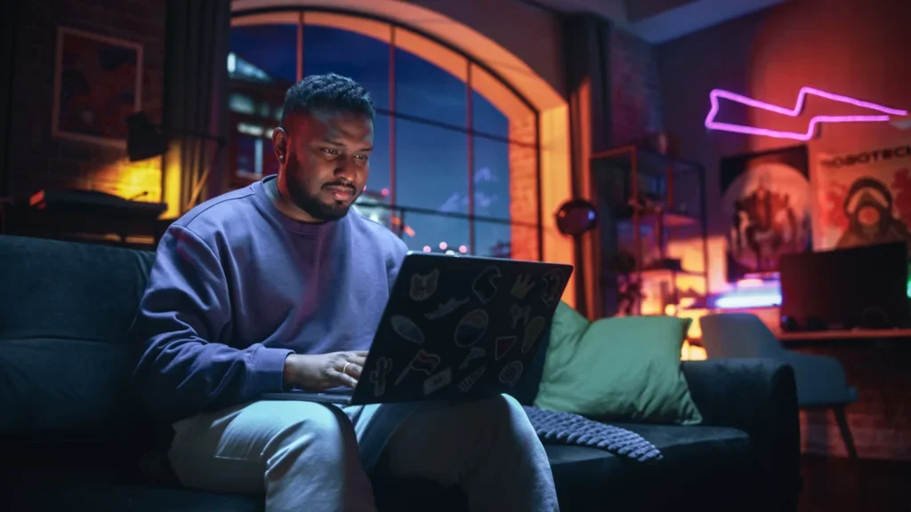 A man sitting on a couch looking at a laptop on his lap, with the laptop back covered in custom stickers.