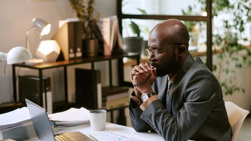 A man in a suit sits at a desk, looking thoughtfully at his laptop with his hands clasped in front of him.