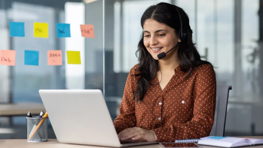 A woman with a headset sits at a desk, focused on her laptop, working as a virtual assistant.