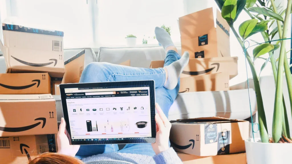 A woman lying on the floor surrounded by Amazon boxes, holding a tablet in front of her with Amazon’s homepage open.