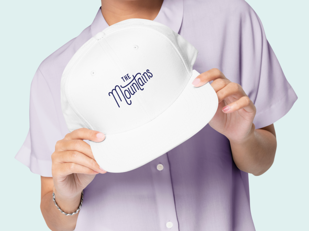 A woman in a light-purple shirt holding a white hat cap with a “The Mountains” print in the center.