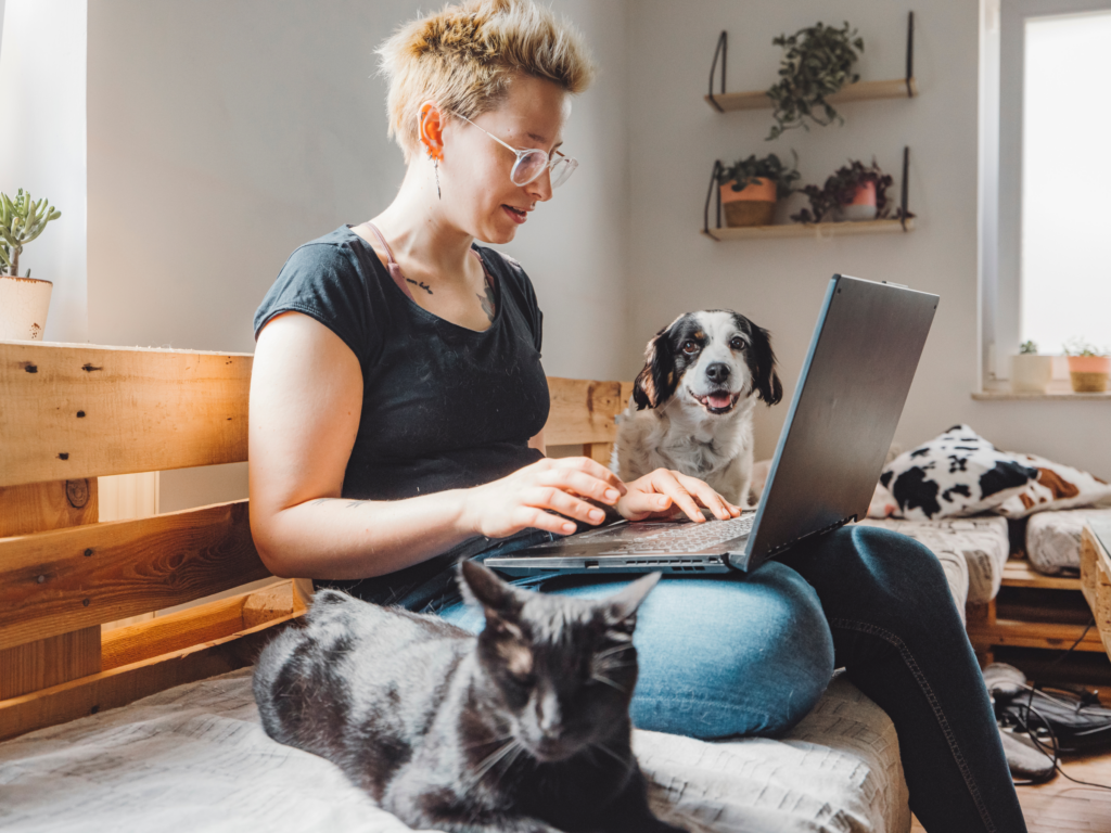 A woman sits on a couch with a laptop, accompanied by a dog and a cat.