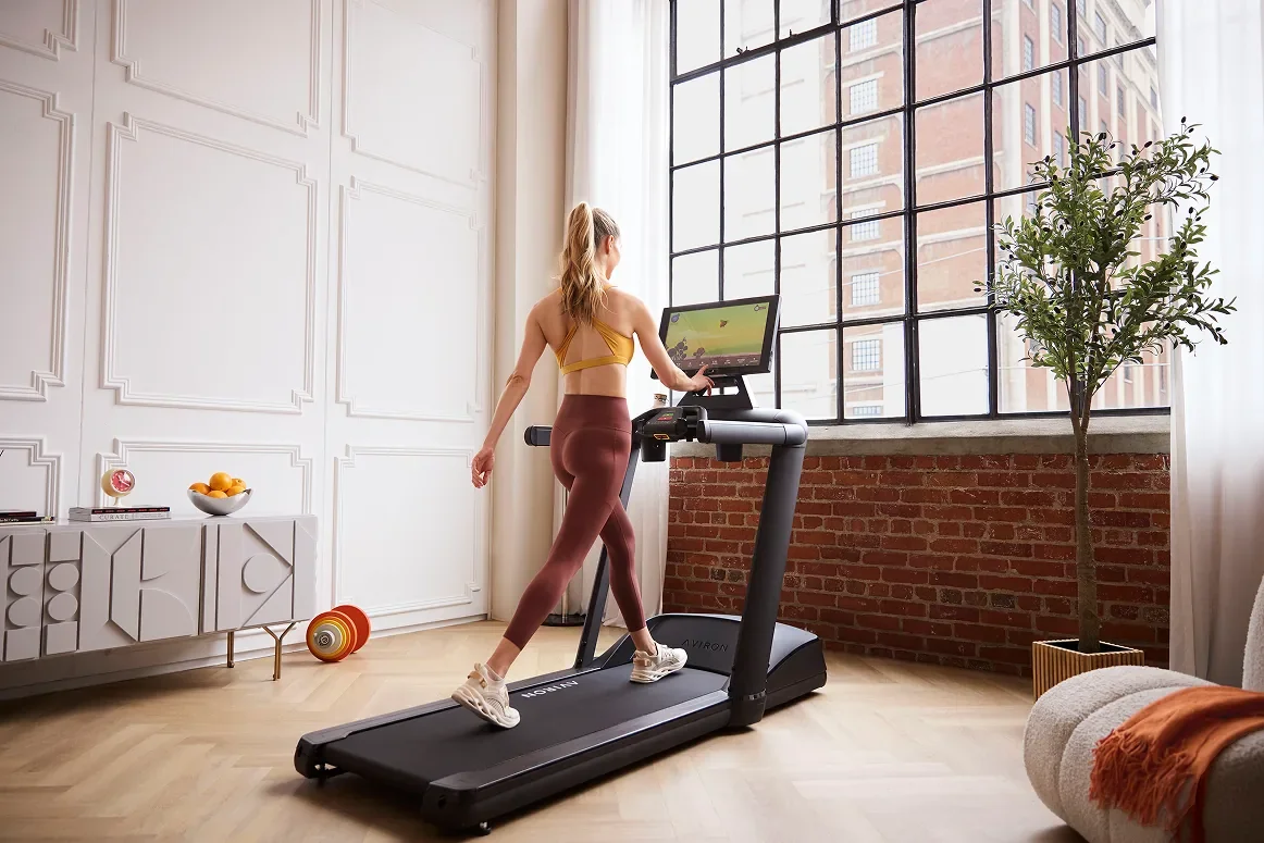 Woman walking on an Aviron Victory treadmill in a bright loft, screen showing a scenic route