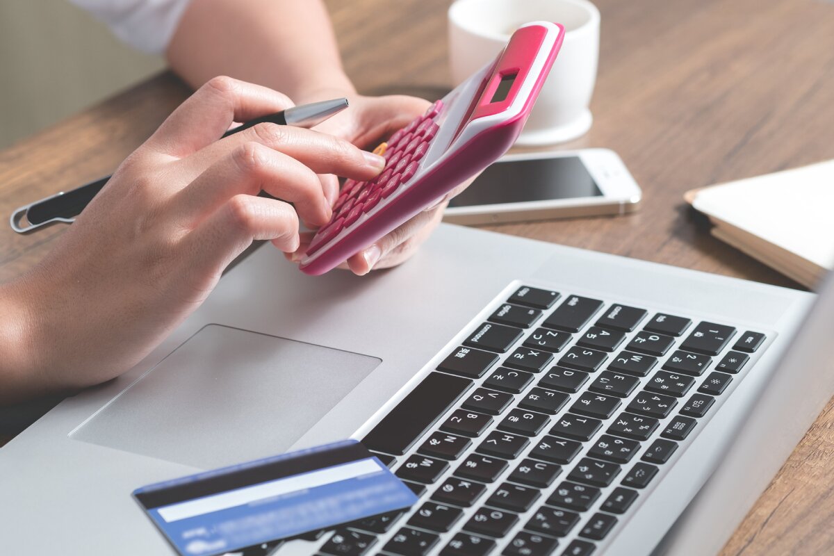 A person sitting at a desk, making calculations with a calculator, a laptop, a mobile phone and a credit card on the surface.