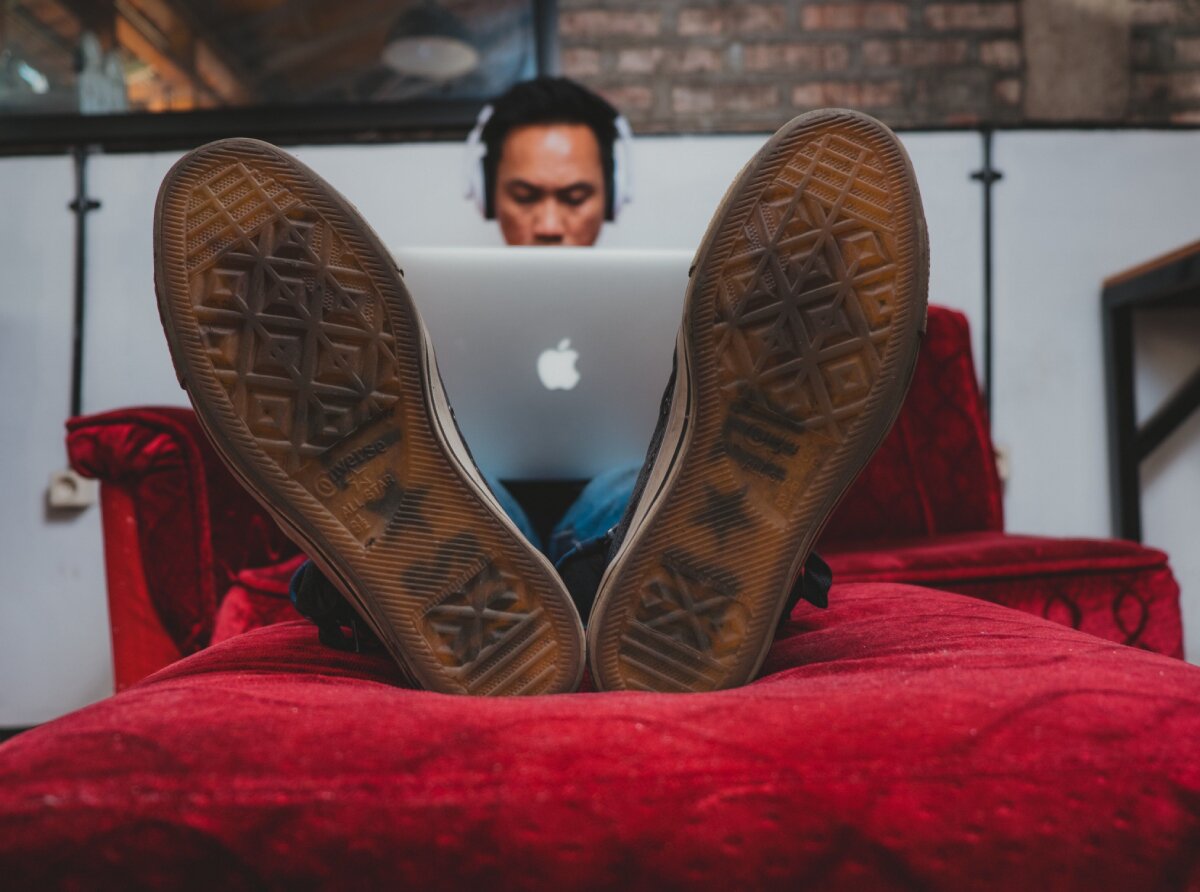 A man sitting with legs resting on a couch, wearing headphones and working on a laptop, viewed from the perspective of his feet