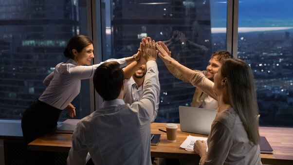 A team celebrates by putting their hands together over a conference room table
