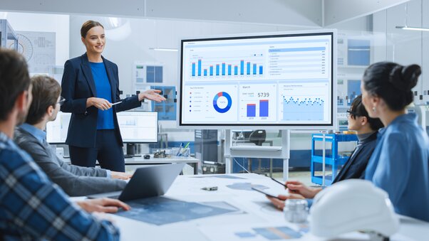 A team member leads an analytics-focused meeting. She gestures toward a screen with charts and graphs. 