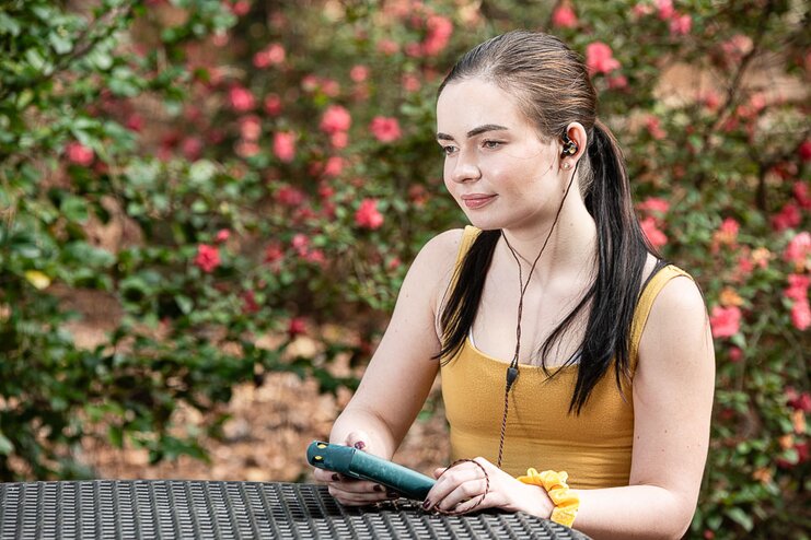 A woman sitting at an outdoor table holding a digital audio player with in-ear monitors on. 