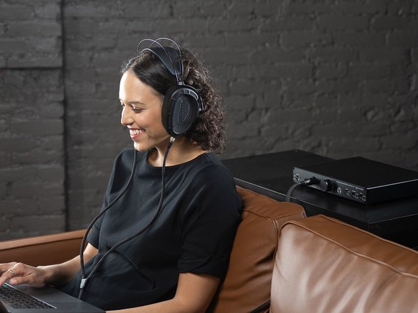 HeadAmp GS-X Mini headphone amplifier lifestyle shot sitting on table behind a woman listening to music with her headphones while using a laptop and sitting on a couch.