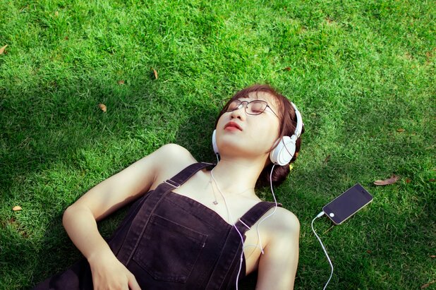 Woman laying on grass with eyes closed wearing headphones connected to a smartphone