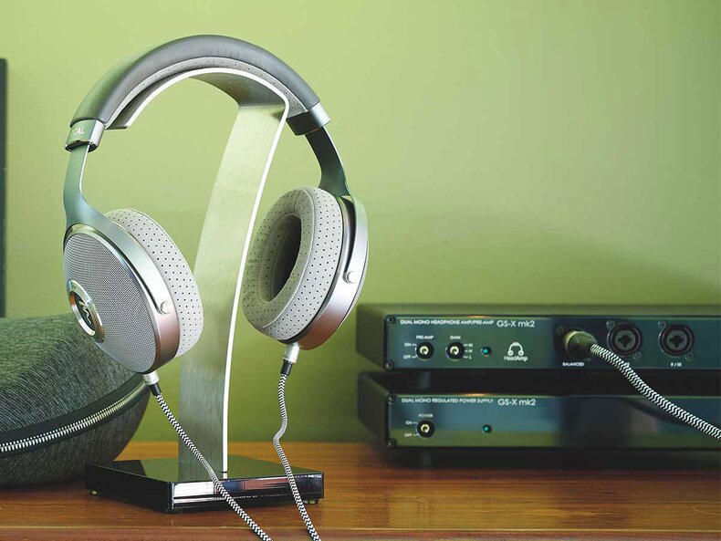 HeadAmp GS-X mk2 Amp sitting on table next to headphones hanging from a stand in front of a green wall.
