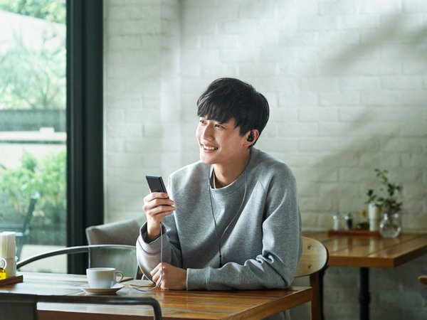 Lifestyle shot of masculine person of Asian decent sitting at a table with a cup of coffee, listening to a Sony Walkman with IEMs.