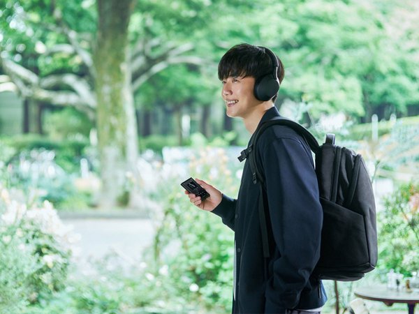 A Masculine person of Asian descent standing outside with a backpack, listening to a Sony Walkman with headphones.