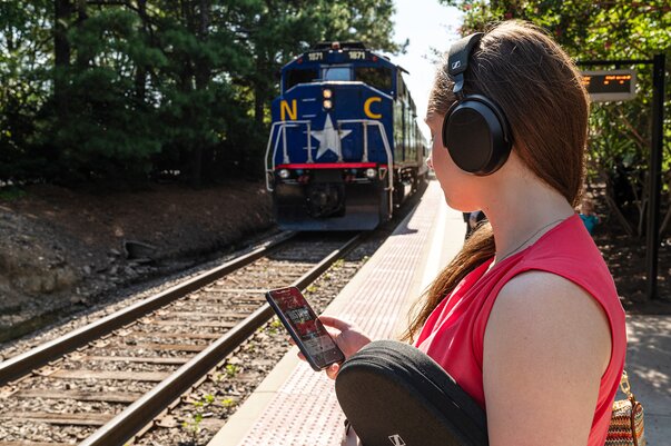 woman waiting for a train on a train platform with a smartphone in one hand, sennheiser momentum headphone carrying case in the other, and sennheiser momentum 4 wireless headphones on. 