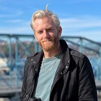 A man with platinum blonde hair and a short beard stands in front of a blue metal bridge under a clear sky. He’s wearing a black jacket over a light blue shirt and a gold chain necklace. He holds a black coffee cup in one hand and looks at the camera with a relaxed smile. The bright daylight and urban background suggest a casual moment captured near a waterfront or scenic overlook.