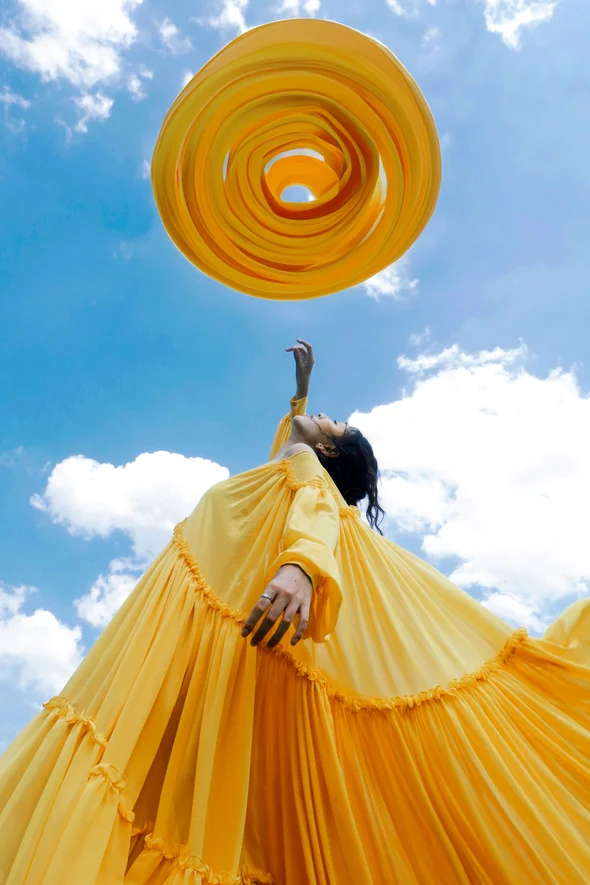 The ESPR: Rethinking how we design clothes. Woman wearing an oversized yellow maxi dress is reaching up to blue skies. 