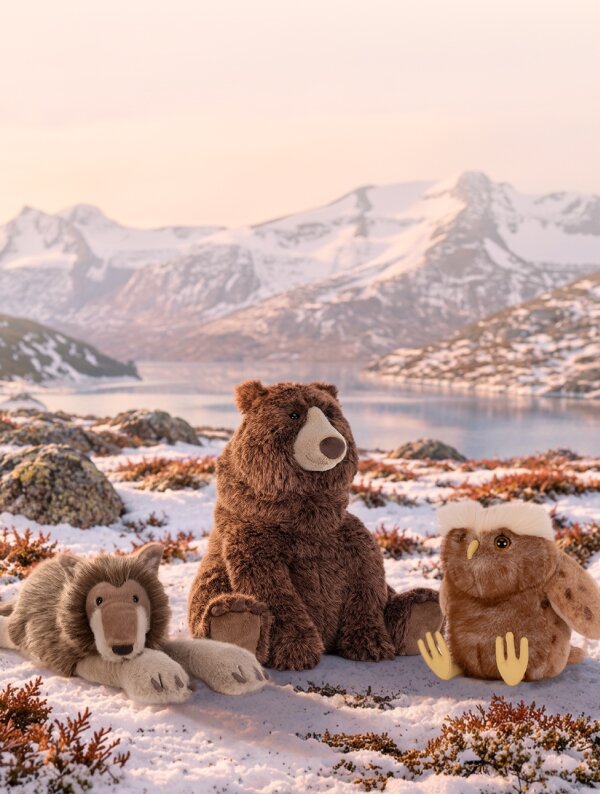 Three plush woodland Jellycats—a wolf, brown bear, and owl—sit together in a snowy tundra landscape with mountains in the background.