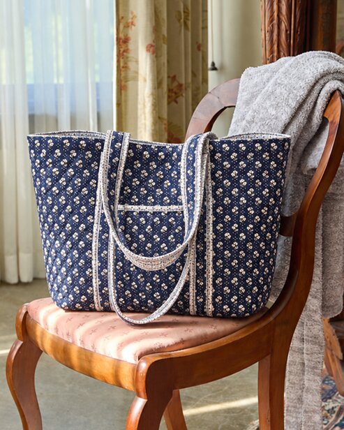 Navy floral quilted Vera Bradley tote bag with patterned trim and double shoulder straps, displayed on a wooden chair beside a cozy gray blanket.