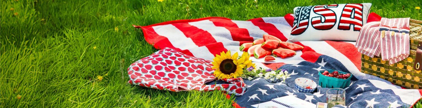 Patriotic picnic setup with an American flag blanket, strawberries, watermelon, flowers, and woven picnic basket spread across green grass.