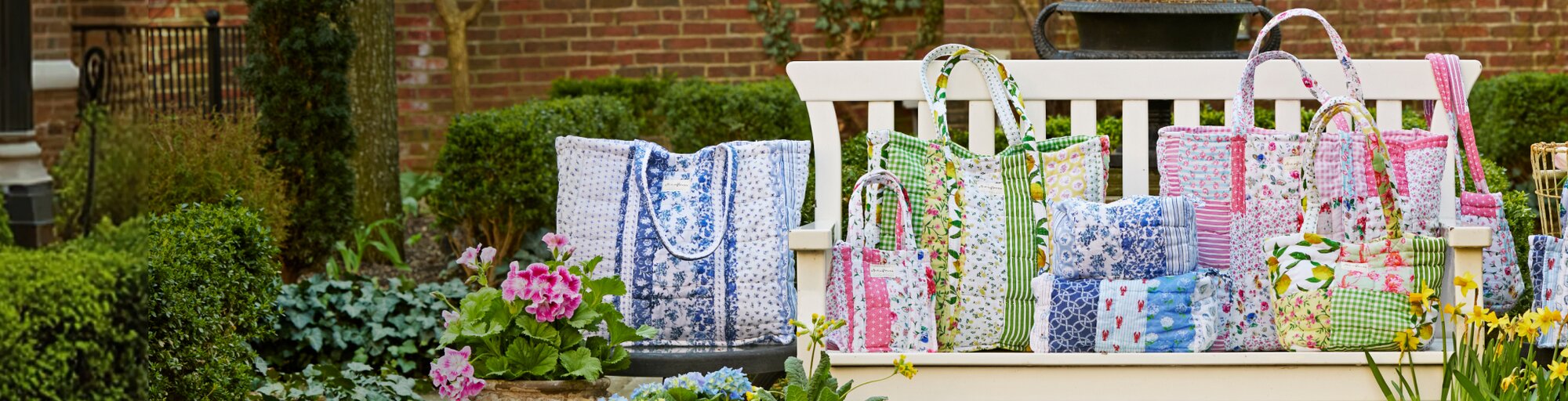 Colorful handmade quilted Strawflower tote bags and cushions in floral and patchwork patterns displayed outdoors on a bench, surrounded by potted flowers and a watering can.