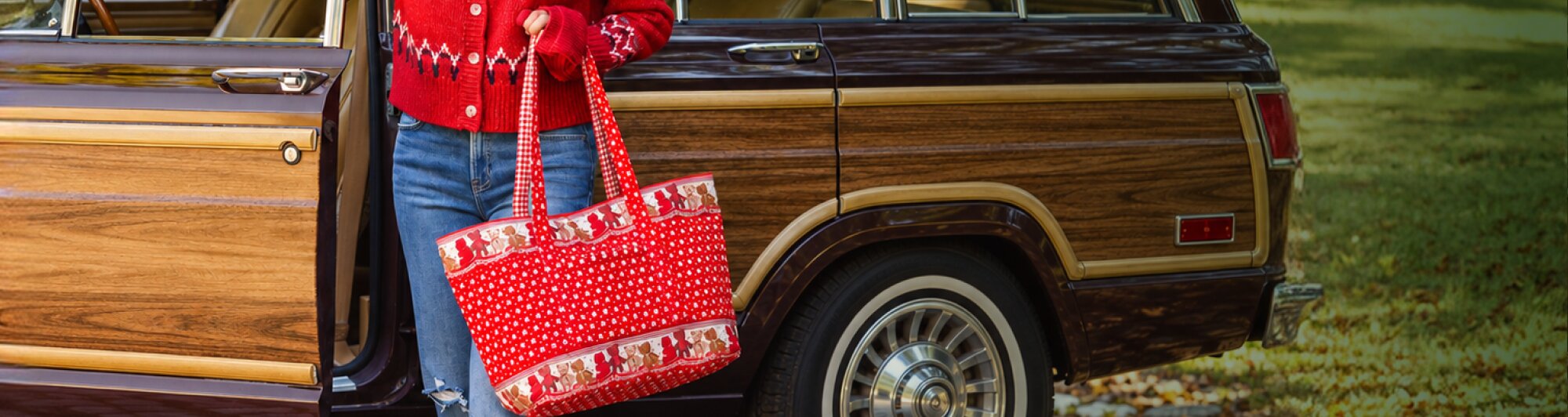 Woman in a red sweater and jeans carrying a red holiday-print Vera Bradley tote bag beside a vintage wood-paneled car.