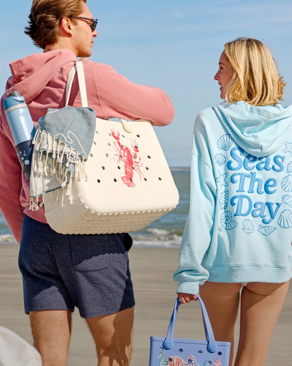 Two people walking along the beach wearing pastel hoodies and carrying a Bogg bag with a towel and Owala water bottle on a sunny day.