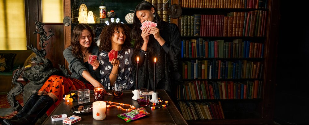 Three people gathered around a table playing cards in a book-lined room decorated with candles, seasonal treats, and Halloween-themed accents.