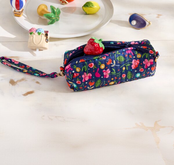 A navy floral zippered Nora Fleming pouch with a wrist strap sits on a light wooden surface, surrounded by small colorful Mini Ornaments like lemon, orange, strawberry,  hot air balloon, with a label indicating it can fit three minis.