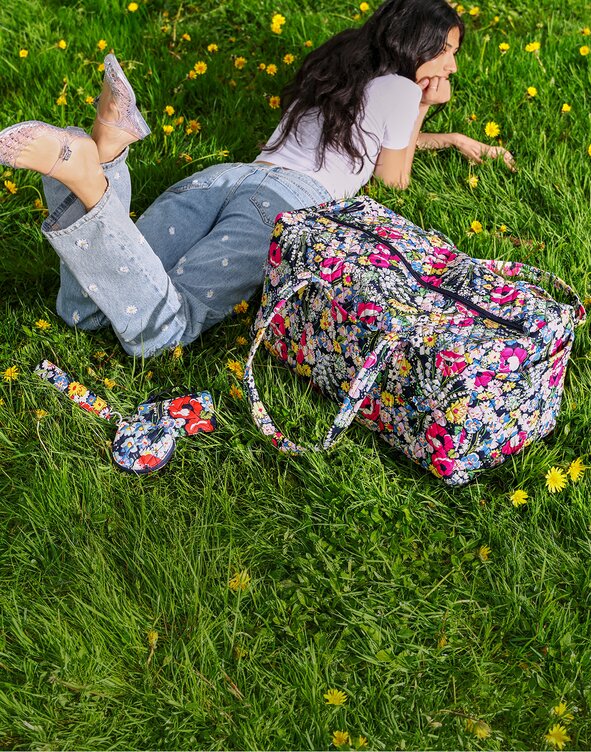 Person lying on green grass dotted with yellow flowers, wearing a white T-shirt, light-wash jeans with embroidered daisies, and clear shoes. A large Vera Bradley duffel bag in Poppy Fields and matching keychain pouch rest beside them, creating a colorful spring-inspired outdoor scene.