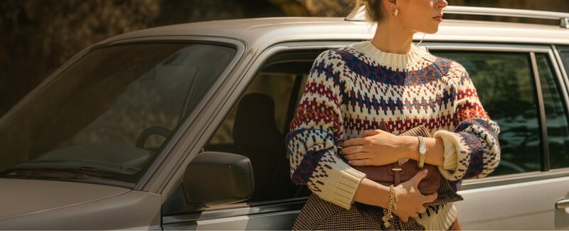 Person standing beside a silver station wagon, wearing a patterned knit sweater and holding a clutch bag in warm outdoor lighting.