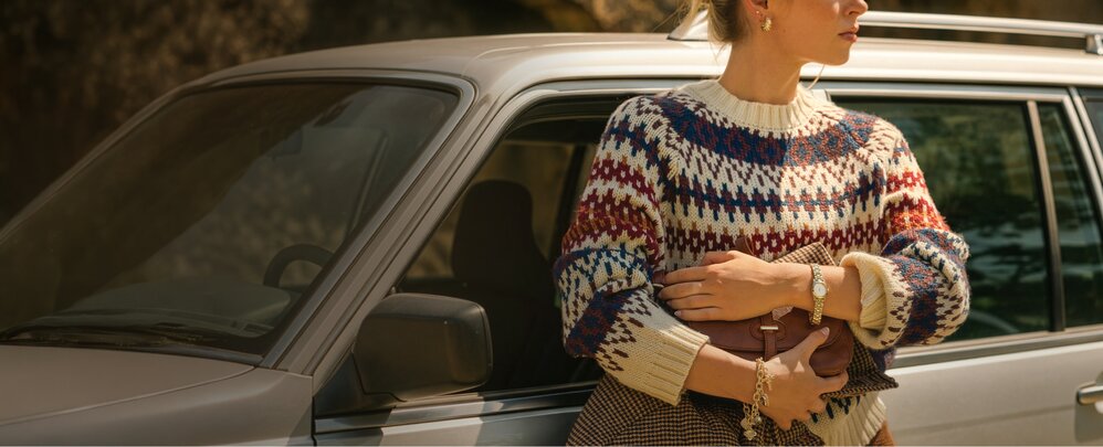 Person standing beside a silver station wagon, wearing a patterned knit sweater and holding a clutch bag in warm outdoor lighting.
