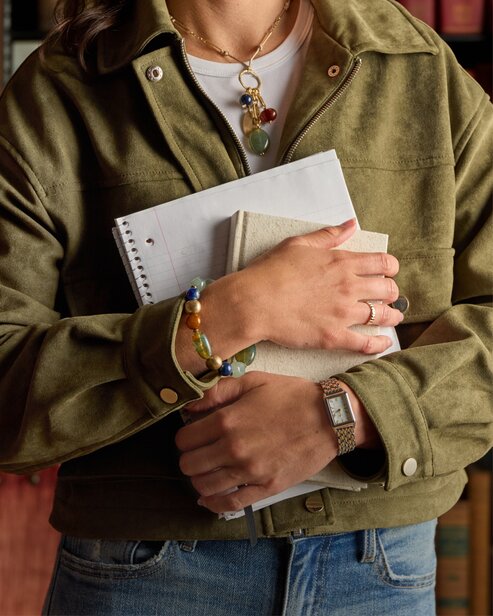 Person holding notebooks while wearing layered necklaces, bracelets, and a green jacket.