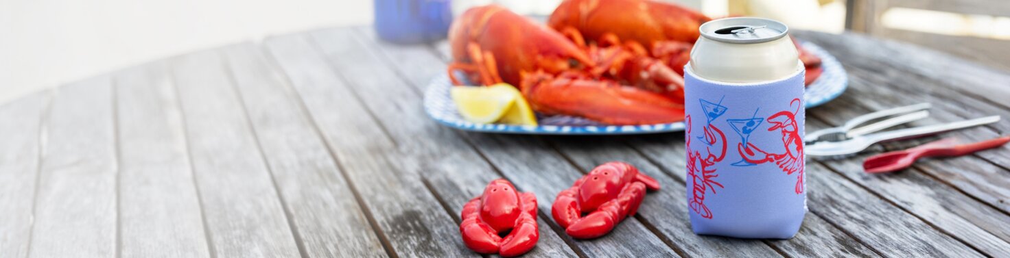 Blue can cooler with red lobster illustrations on a weathered picnic table beside lobster-themed salt & pepper shakers and seafood.
