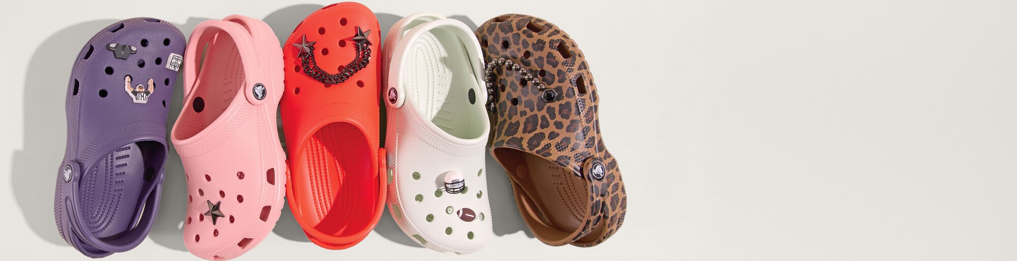 A row of five colorful Crocs clogs arranged side by side on a light background. Each shoe features decorative charms: a purple pair with small icons, a pink pair with a star, a bright red pair with star and chain accents, a pale green pair with simple charms, and a brown pair with a leopard print pattern.